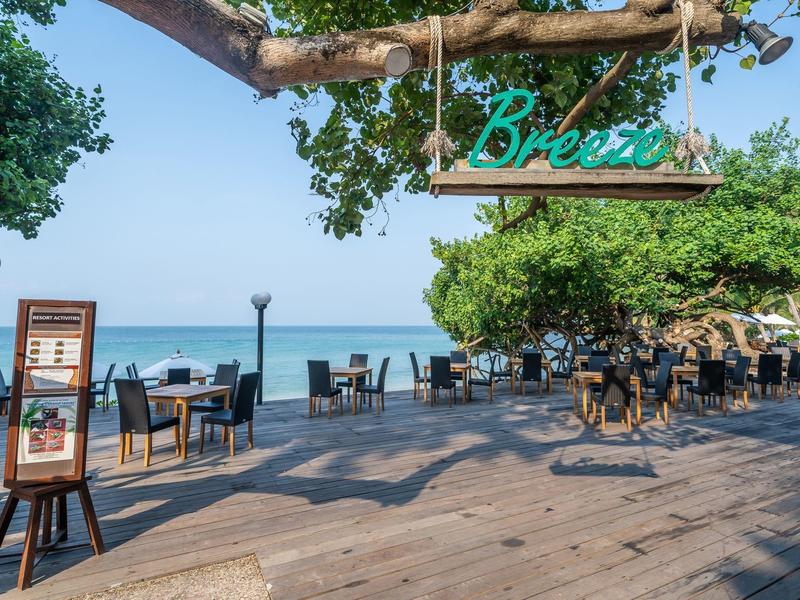 Freiluft-Café mit Holzboden, Tischen, Stühlen und Blick auf das blaue Meer bei klarem Himmel.