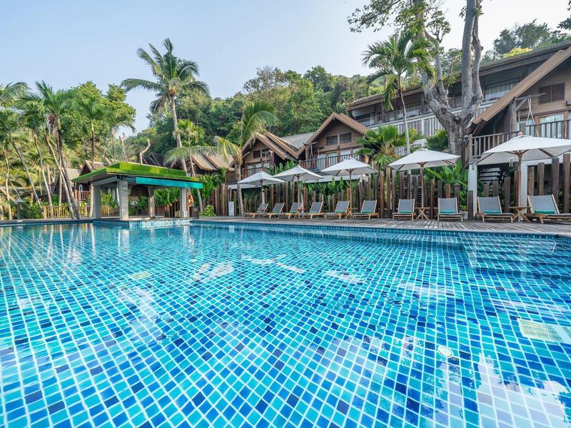 Großer Pool mit blauen Fliesen, umgeben von Holzbungalows und grünen Bäumen im Hintergrund.