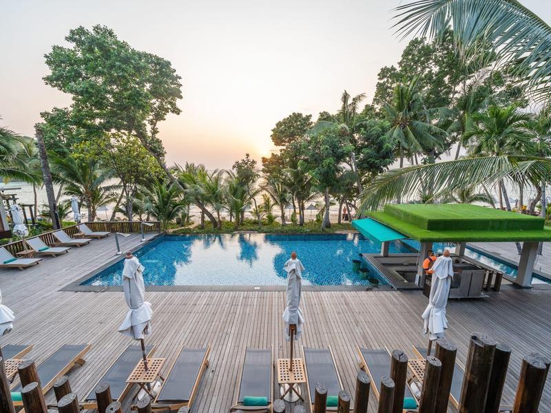 Großer Swimmingpool mit Holzdeck, Sonnenliegen, Palmen und Blick auf das Meer bei Sonnenuntergang.