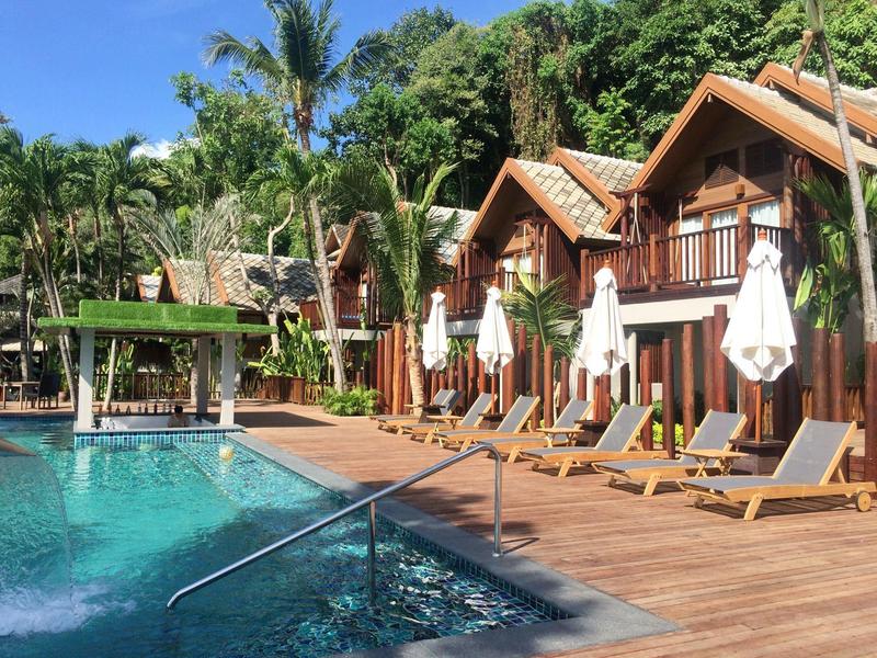 Pool mit Liegestühlen und Sonnenschirmen vor Holzhäusern, Palmen und bewaldetem Hintergrund.