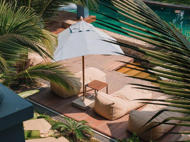 Gemütliche Poolterrasse mit Holzboden, Sonnenschirm, Sitzkissen und Palmenblättern im Vordergrund.