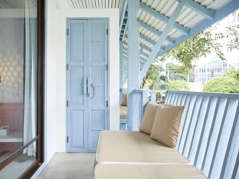 Blauer Balkon mit Sofa, Kissen, Holztür und Blick auf Bäume und Gebäude im Hintergrund.