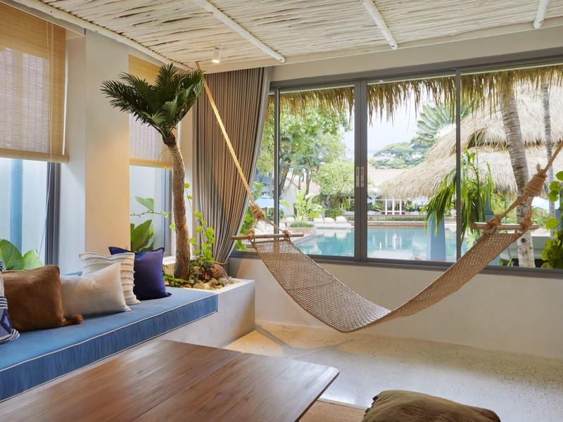 Helle Wohnzimmer mit hängender Hängematte, blauen Sitzkissen und großer Fensterfront mit Palmenblick.