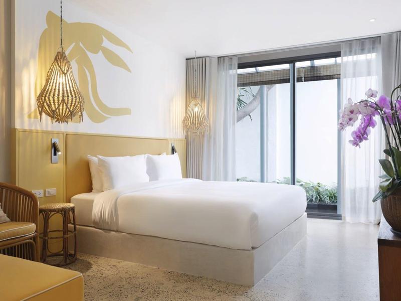 Modernes Schlafzimmer mit Doppelbett, Holzmöbeln, Korbsessel und großen Fenstern mit Gardinen.