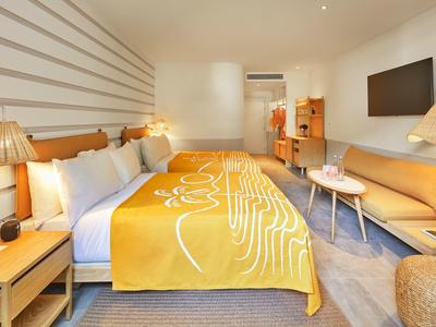 Modernes Hotelzimmer mit Bett, orangefarbener Decke, Sofa, Fernseher und warmem Licht.