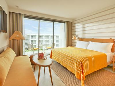 Helles Hotelzimmer mit großem Fenster, orangefarbenem Sofa, Bett mit oranger Bettdecke und Holzmöbeln.