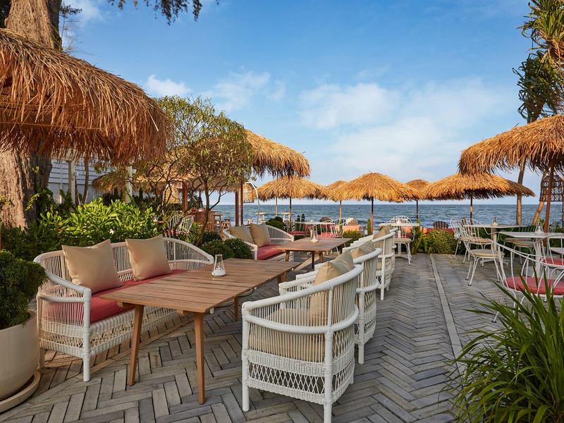 Freiluftrestaurant mit weißen Sesseln, Holztischen und Strohdächern bei klarem Himmel am Meer.