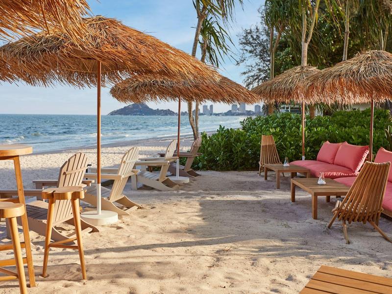 Strand mit Sand, Strohhut-Sonnenschirmen, Holzstühlen und gemütlichen Sitzmöbeln am Meer.