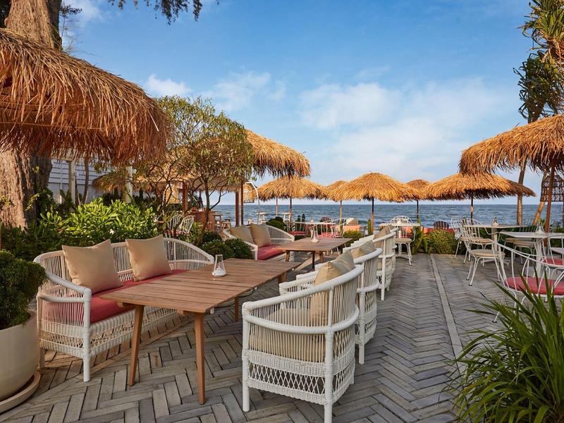 Außenbereich eines Cafés mit weißen Stühlen, Holztischen und Strohdächern am Meer bei blauem Himmel.