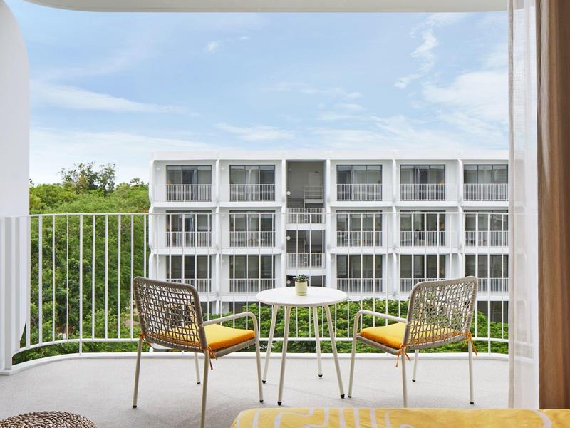 Balkon mit zwei Stühlen und kleinem Tisch, moderner weißer Gebäudekomplex im Hintergrund.