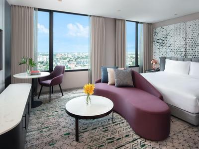 Habitación de hotel moderna con ventana grande, sofá morado, cama blanca y mesa pequeña con flor.