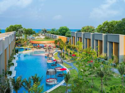 Complejo hotelero moderno con gran piscina, vegetación tropical y vista al mar de fondo.