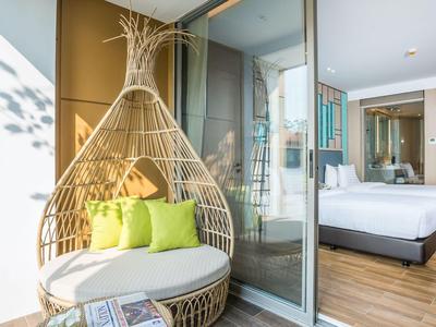 Balcón moderno de hotel con silla de mimbre y vista a una habitación iluminada con cama doble.