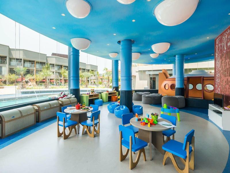 Espace intérieur bleu clair avec tables et chaises pour enfants près de la piscine et grandes fenêtres.