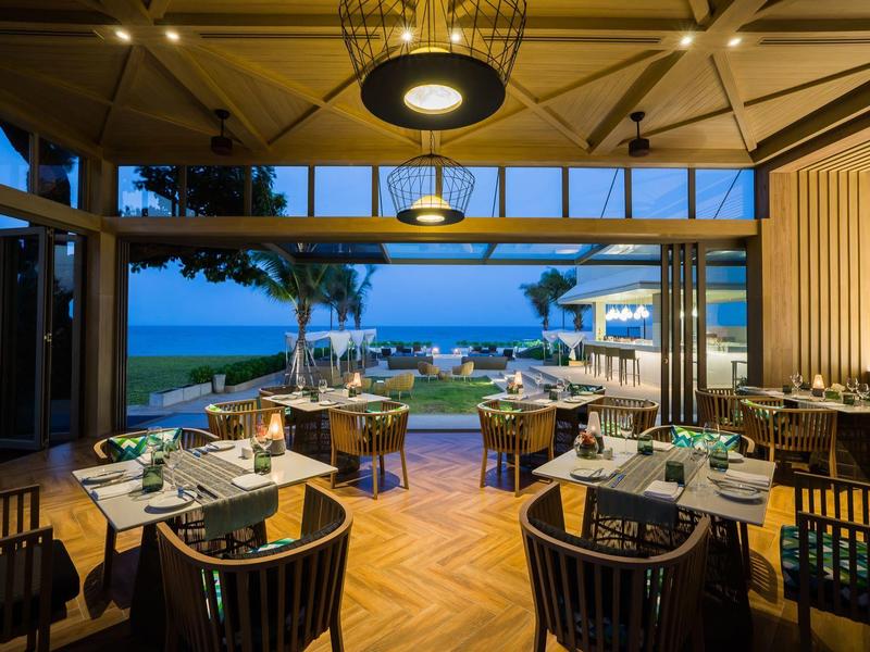 Restaurant ouvert avec vue sur la plage, ambiance du soir avec tables élégamment dressées et parquet en bois.