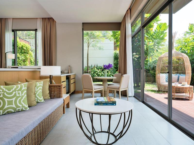 Salon lumineux avec canapé, coussins et table devant de grandes fenêtres donnant sur une terrasse avec sièges.