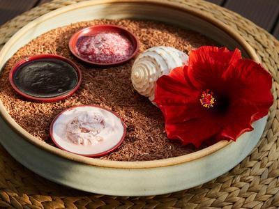 Miska s pískem, mušlí, červeným květem a malými nádobkami s krémy na pleteném povrchu.