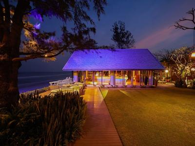 Osvětlený dřevěný dům u moře při západu slunce s zahradou a stromy.