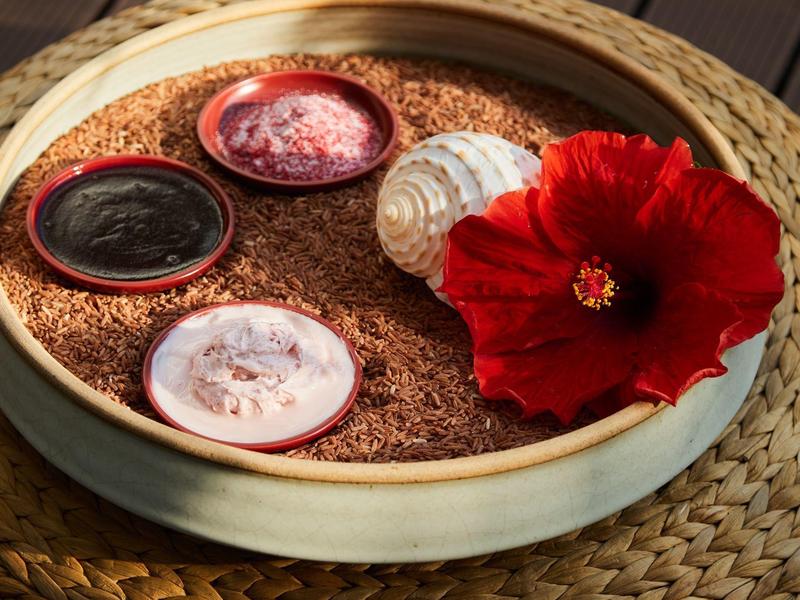 Miska s pískem, mušlí, červeným květem a malými nádobkami s krémy na pleteném povrchu.