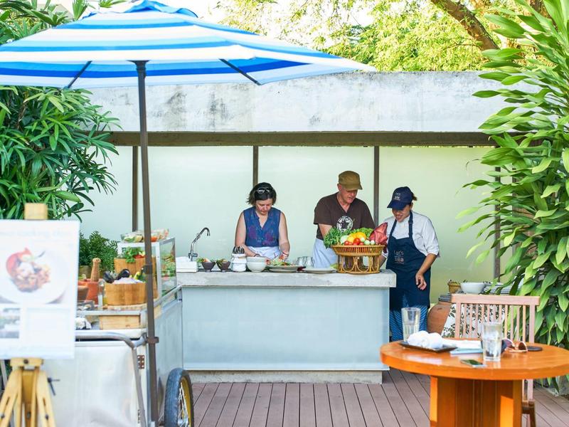 Tři lidé připravují jídlo u venkovního bufetu pod modrým slunečníkem.