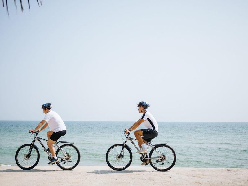 Dva lidé jedou na kolech po pláži vedle moře pod jasnou oblohou.