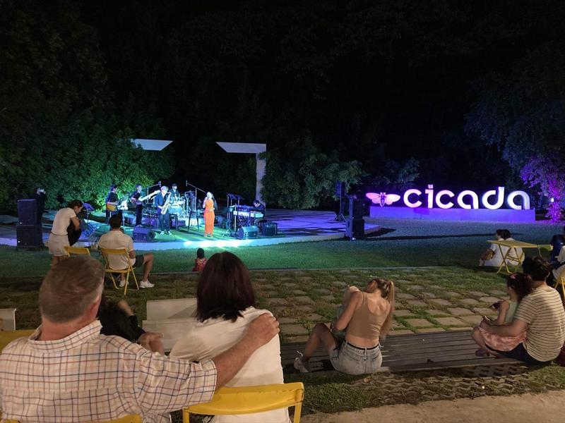 Noční scéna venkovního koncertu s publikem sedícím na schodech a osvětleným nápisem 'cicada'