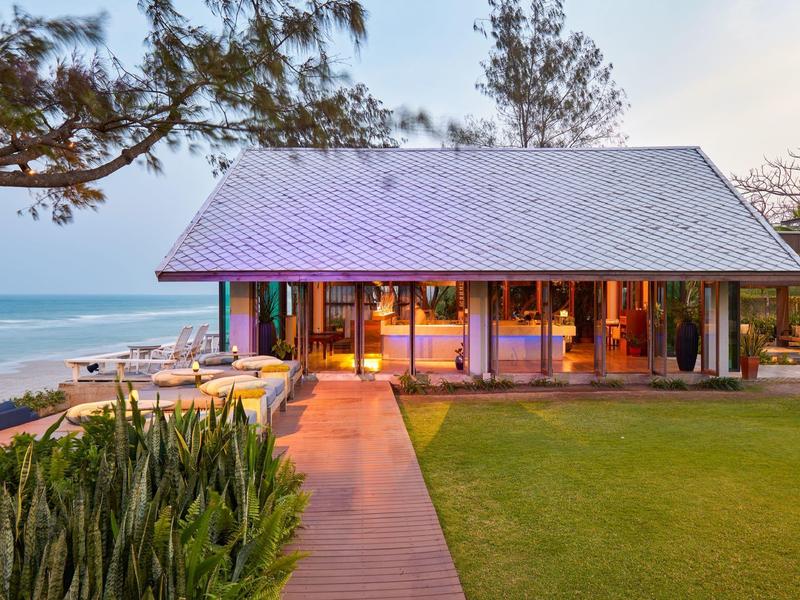 Moderní dům s dřevěnou cestou u pláže a tropickou zahradou při západu slunce.