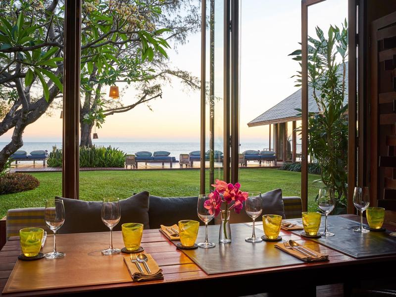 Stůl se sklenicemi a květinami v restauraci s výhledem na zahradu a moře.