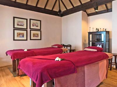 Spokojny pokój do masażu z dwoma stołami, czerwonymi pokrowcami i dekoracyjnymi obrazami na ścianie.