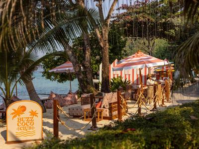 Strandrestaurant mit Tischen und Stühlen unter Bäumen am Meer, umgeben von Palmen.