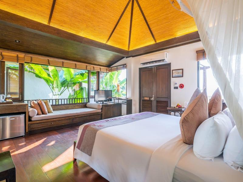 Jasny pokój hotelowy z dużym łóżkiem, miejscem do siedzenia, drewnianym sufitem i widokiem na rośliny tropikalne.