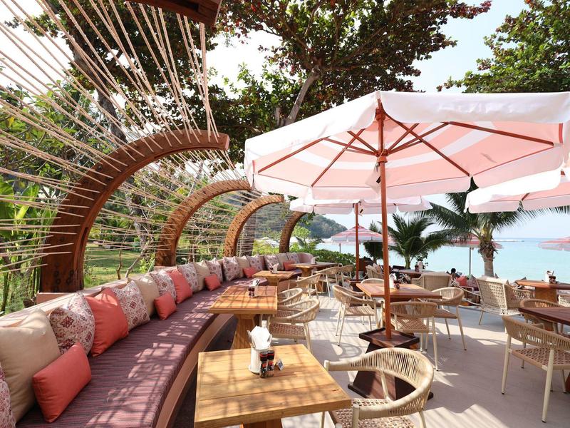 Przytulny salon z drewnianymi siedzeniami, poduszkami i parasolami na tle tropikalnej plaży.