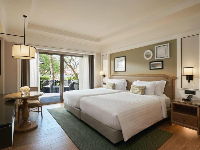 Bright hotel room with two double beds, light fixtures, and balcony overlooking greenery.