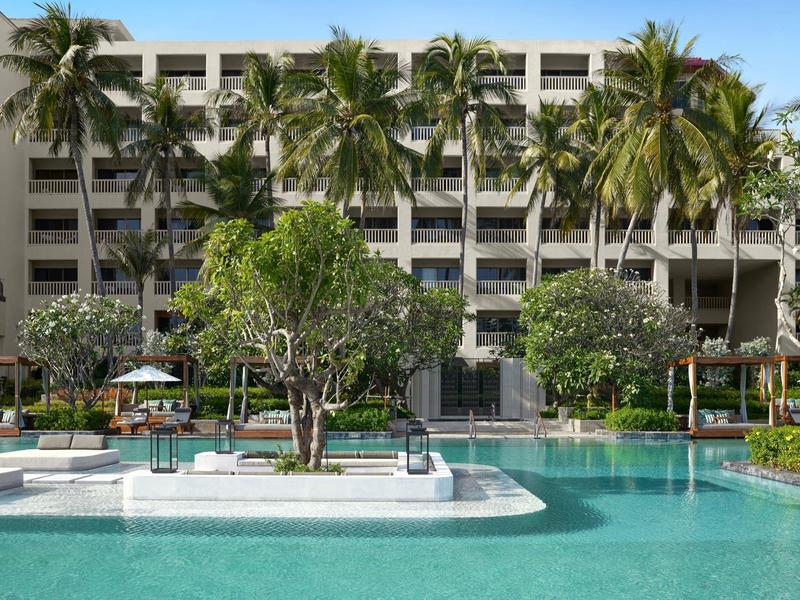 Large outdoor pool with island and palm trees in front of a multi-story hotel building under clear skies.