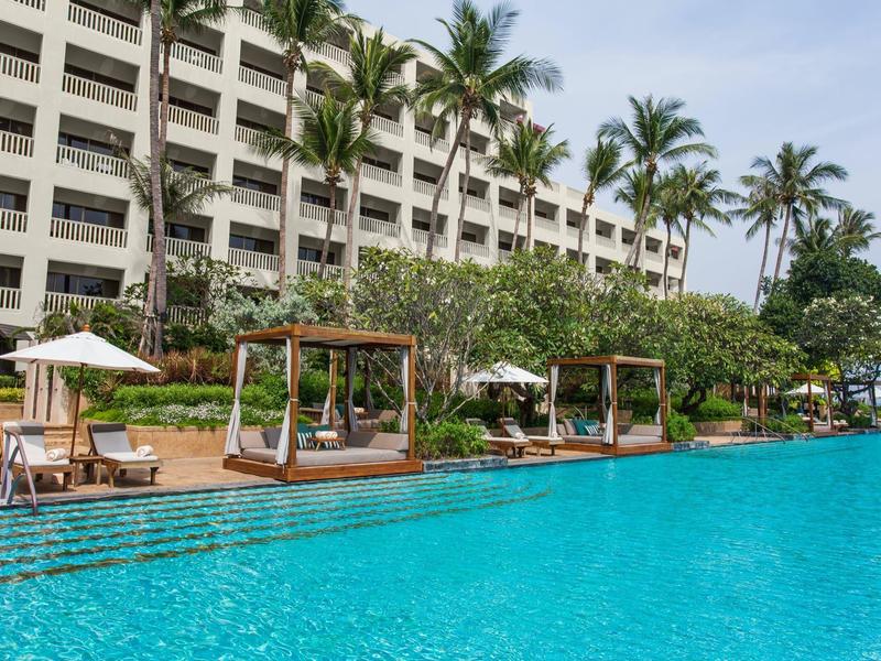 Large pool with daybeds and palm trees in front of a multi-story hotel building.
