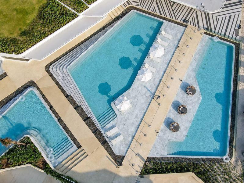 Vista aerea di una piscina moderna con lettini e ombrelloni accanto a un hotel.