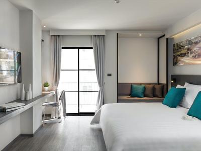 Modern hotel room with double bed, TV, desk, and balcony with bright windows.