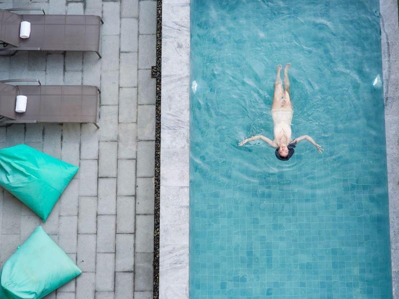 Person floats on their back in a narrow outdoor pool beside lounge chairs.