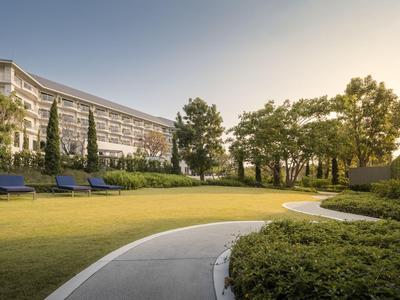 Spacious hotel grounds with green lawn, seating areas, and pathways in sunlight.