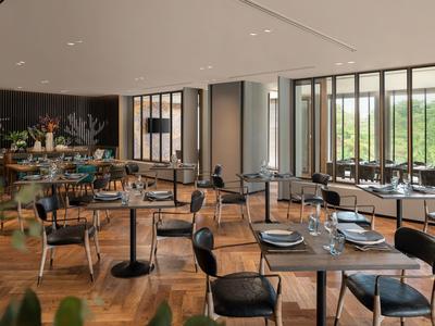 Modern restaurant with wooden floor, black chairs, and large windows with views of greenery.