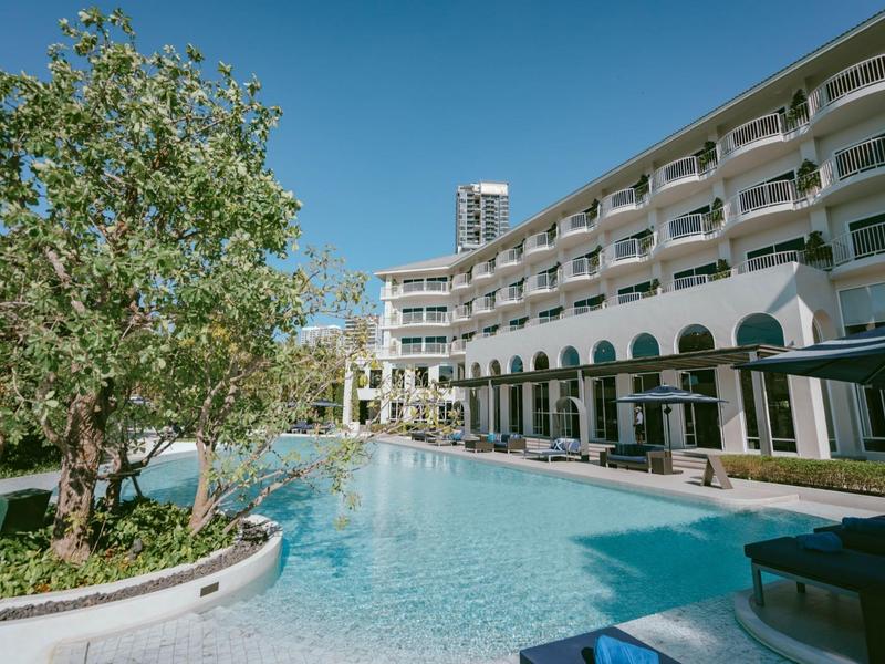 Hotel building with outdoor pool, surrounded by lounge chairs and trees under a clear sky.