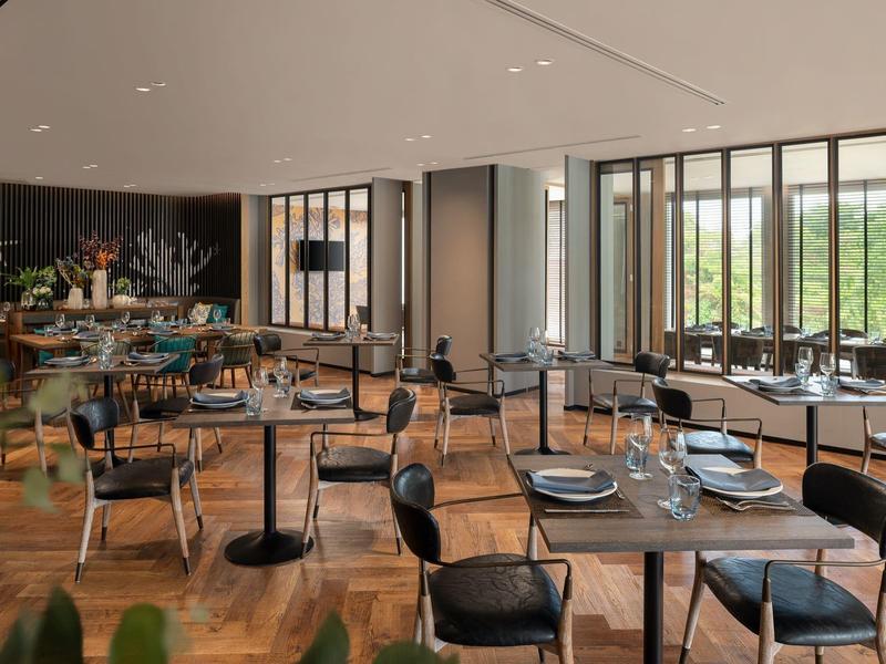 Modern restaurant with wooden floor, black chairs, and large windows with views of greenery.