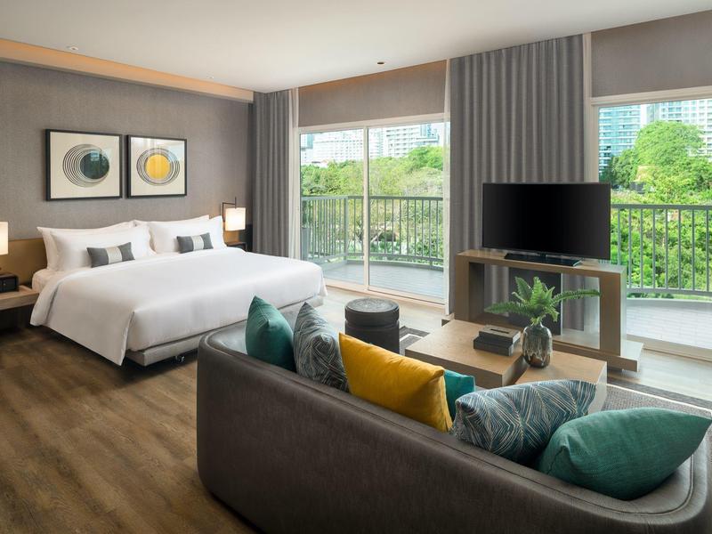 Modern hotel room with king-size bed, sofa, TV, and balcony overlooking greenery.