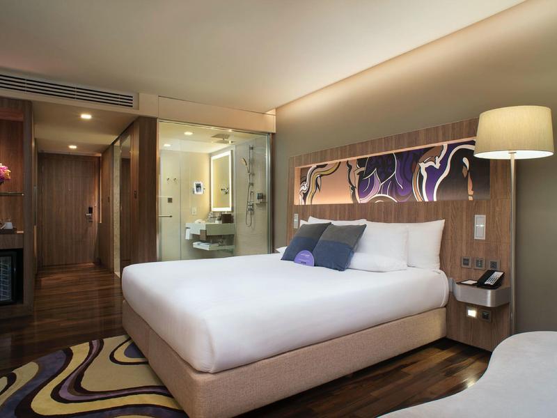 Habitación de hotel moderna con cama grande, iluminación indirecta y baño con pared de vidrio.