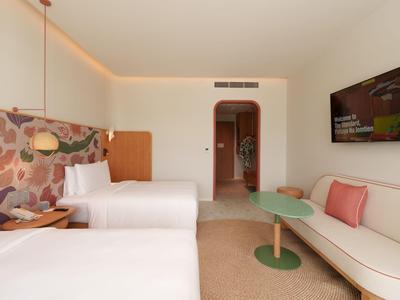 Helles Hotelzimmer mit zwei Betten, floraler Wanddeko, Sofa und modernem Minimaldesign.