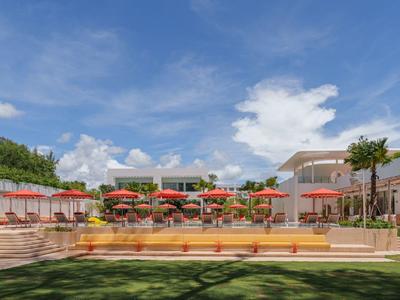 Außenansicht eines Hotels mit Garten, Terrasse und bunten Sonnenschirmen unter blauem Himmel.