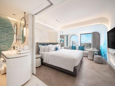 Modernes Hotelzimmer mit großem Bett, Sofa, Fernseher und stilvollem Design in hellen Farben.