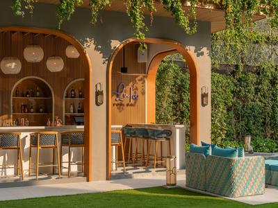 Moderne Hotelbar mit Holztheke, Bistromöbeln und grüner Gartenkulisse bei Sonnenlicht.