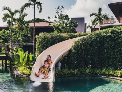 Two people slide down a water slide into a pool surrounded by tropical plants.