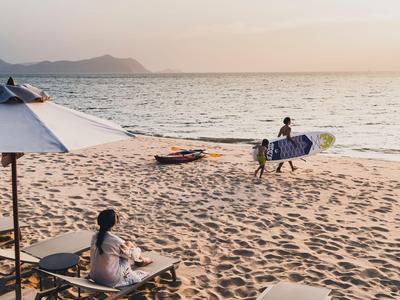 A person carries a surfboard on the beach at sunset, with people sitting under a sunshade in the foreground.